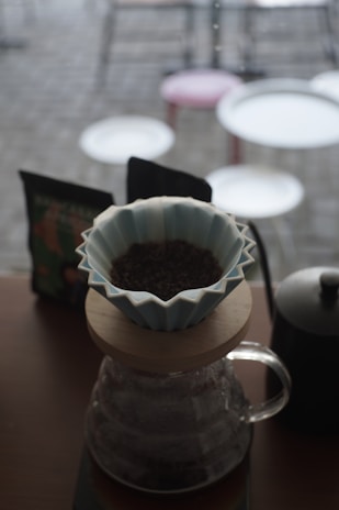 A coffee brewing setup with a ceramic dripper placed on top of a glass carafe. Ground coffee is visible in the dripper. In the background, there are blurred images of tables and chairs, suggesting a cafe environment.