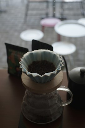 A coffee brewing setup with a ceramic dripper placed on top of a glass carafe. Ground coffee is visible in the dripper. In the background, there are blurred images of tables and chairs, suggesting a cafe environment.