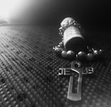 A rolled-up piece of paper is surrounded by a beaded rosary with a cross. The cross features the word 'Jesus'. The surface beneath has a textured, fabric-like pattern, and the image has a monochromatic appearance with a high contrast between light and dark areas.