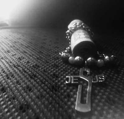 A rolled-up piece of paper is surrounded by a beaded rosary with a cross. The cross features the word 'Jesus'. The surface beneath has a textured, fabric-like pattern, and the image has a monochromatic appearance with a high contrast between light and dark areas.