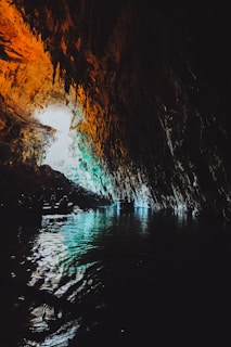 A cavern with a glowing opening is illuminated by natural light, revealing the textured rock formations with orange and cyan hues. The dark, reflective water below carries a boat with several silhouetted figures, creating a tranquil and mysterious atmosphere.