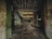An abandoned industrial building with graffitied walls and mold covering many surfaces. The ceiling shows signs of decay with peeling material, and the floor is littered with debris and puddles. Light filters through openings, highlighting the dilapidation.