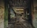 An abandoned industrial building with graffitied walls and mold covering many surfaces. The ceiling shows signs of decay with peeling material, and the floor is littered with debris and puddles. Light filters through openings, highlighting the dilapidation.