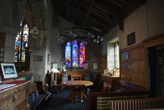 A dimly lit interior of a church or chapel featuring intricate stained glass windows depicting religious scenes. The wooden furniture includes pews and a central table surrounded by chairs with ornate designs. The stone walls lend a historic and solemn atmosphere. Various framed pictures and plaques are displayed along the walls.