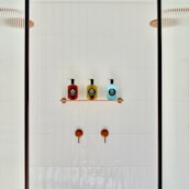 A minimalist shower area featuring a white tiled wall with two copper shower controls. A small shelf holds three soap dispensers in red, yellow, and blue, each with black labels. Partial glass panels are visible on either side.