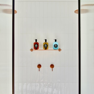 A minimalist shower area featuring a white tiled wall with two copper shower controls. A small shelf holds three soap dispensers in red, yellow, and blue, each with black labels. Partial glass panels are visible on either side.