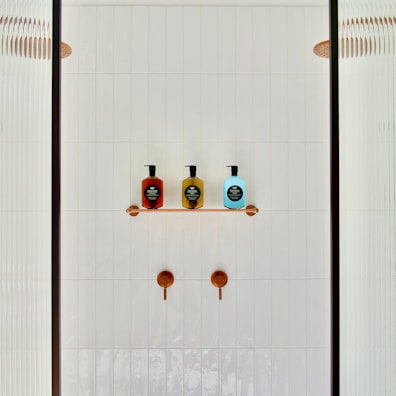 A minimalist shower area featuring a white tiled wall with two copper shower controls. A small shelf holds three soap dispensers in red, yellow, and blue, each with black labels. Partial glass panels are visible on either side.