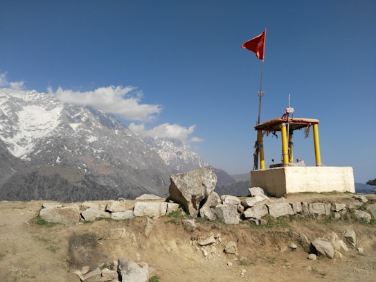 A small shrine with yellow pillars and a red flag is situated on a rocky outcrop. In the background, snow-capped mountains extend across the horizon under a clear blue sky with a few clouds.