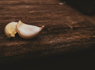 Two cloves of garlic are resting on a rustic wooden surface. The wood has a rough texture, with visible grain and marks, adding an earthy and natural backdrop to the scene.