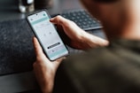 A person is holding a smartphone in their hand, focusing on a timer or scheduling app displayed on the screen. The person's arm and part of the torso are visible. The setting appears to be a workspace with a keyboard slightly out of focus in the background.