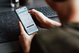 A person is holding a smartphone in their hand, focusing on a timer or scheduling app displayed on the screen. The person's arm and part of the torso are visible. The setting appears to be a workspace with a keyboard slightly out of focus in the background.