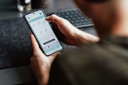 A person is holding a smartphone in their hand, focusing on a timer or scheduling app displayed on the screen. The person's arm and part of the torso are visible. The setting appears to be a workspace with a keyboard slightly out of focus in the background.