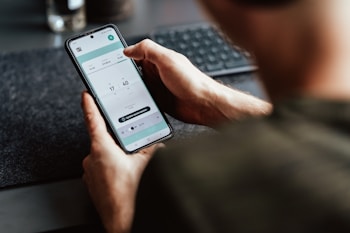 A person is holding a smartphone in their hand, focusing on a timer or scheduling app displayed on the screen. The person's arm and part of the torso are visible. The setting appears to be a workspace with a keyboard slightly out of focus in the background.