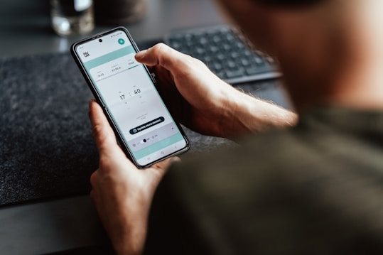 A person is holding a smartphone in their hand, focusing on a timer or scheduling app displayed on the screen. The person's arm and part of the torso are visible. The setting appears to be a workspace with a keyboard slightly out of focus in the background.