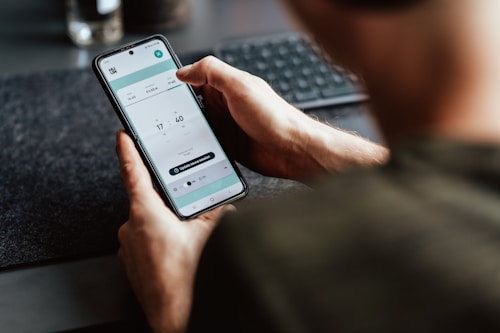 A person is holding a smartphone in their hand, focusing on a timer or scheduling app displayed on the screen. The person's arm and part of the torso are visible. The setting appears to be a workspace with a keyboard slightly out of focus in the background.