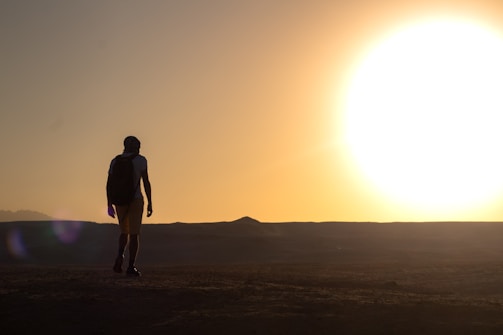 A silhouette of a person with a backpack walking on a barren landscape with a bright sun setting on the horizon, casting a golden glow across the scene.