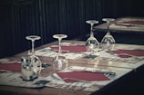 A restaurant table is set for a meal with four wine glasses placed upside down, red napkins, cutlery, and menus laid out for four settings. The ambiance is dimly lit, suggesting a cozy dining atmosphere.