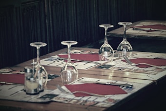 A restaurant table is set for a meal with four wine glasses placed upside down, red napkins, cutlery, and menus laid out for four settings. The ambiance is dimly lit, suggesting a cozy dining atmosphere.