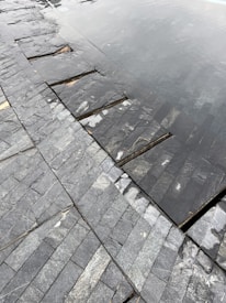 A close-up view of a tiled surface with dark, slate-like stones arranged in a neat pattern. Part of the surface is submerged under shallow water, creating a reflective effect. The textures of the stones are varied, with some showing light veining and natural blemishes.