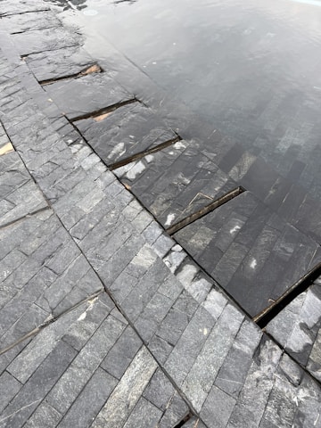 A close-up view of a tiled surface with dark, slate-like stones arranged in a neat pattern. Part of the surface is submerged under shallow water, creating a reflective effect. The textures of the stones are varied, with some showing light veining and natural blemishes.