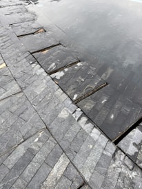 A close-up view of a tiled surface with dark, slate-like stones arranged in a neat pattern. Part of the surface is submerged under shallow water, creating a reflective effect. The textures of the stones are varied, with some showing light veining and natural blemishes.