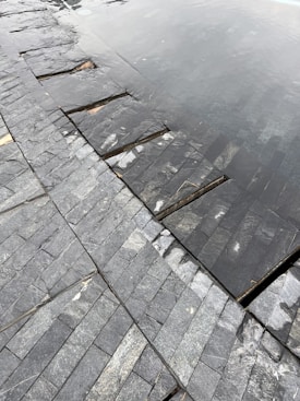 A close-up view of a tiled surface with dark, slate-like stones arranged in a neat pattern. Part of the surface is submerged under shallow water, creating a reflective effect. The textures of the stones are varied, with some showing light veining and natural blemishes.