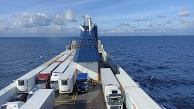 A large ferry deck carrying several trucks, surrounded by a vast ocean under a blue sky with scattered clouds. The ship's structure is visible, with a central chimney and various container trucks lined up on both sides.