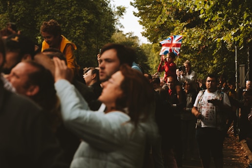 A crowd of people gathered outdoors, with some individuals capturing moments using cameras or phones. A child sitting on someone's shoulders is holding a Union Jack flag, which adds to the vibrant atmosphere. The scene is surrounded by green trees, suggesting a park setting.