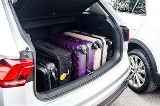 A car's trunk filled with multiple suitcases, including a black, a purple, and a beige one, each with wheels and handles visible. The trunk is open, revealing the neatly packed luggage, and part of another white car is visible beside it.