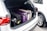 A car's trunk filled with multiple suitcases, including a black, a purple, and a beige one, each with wheels and handles visible. The trunk is open, revealing the neatly packed luggage, and part of another white car is visible beside it.
