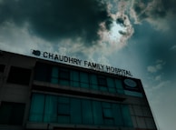 A hospital building with a sign that reads 'Chaudhry Family Hospital', positioned under a cloudy sky with visible sunlight peeking through. The building has multiple windows, some of which are covered or tinted.