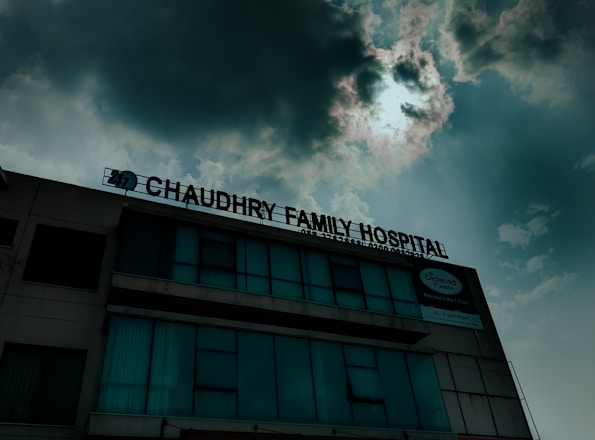A hospital building with a sign that reads 'Chaudhry Family Hospital', positioned under a cloudy sky with visible sunlight peeking through. The building has multiple windows, some of which are covered or tinted.