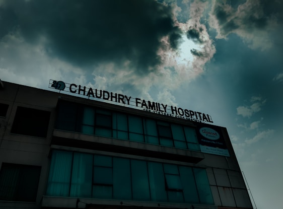 A hospital building with a sign that reads 'Chaudhry Family Hospital', positioned under a cloudy sky with visible sunlight peeking through. The building has multiple windows, some of which are covered or tinted.