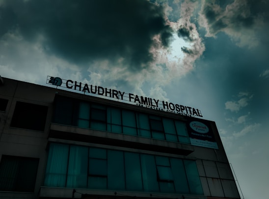 A hospital building with a sign that reads 'Chaudhry Family Hospital', positioned under a cloudy sky with visible sunlight peeking through. The building has multiple windows, some of which are covered or tinted.