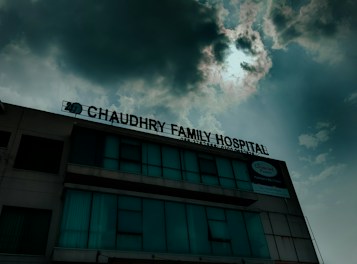 A hospital building with a sign that reads 'Chaudhry Family Hospital', positioned under a cloudy sky with visible sunlight peeking through. The building has multiple windows, some of which are covered or tinted.