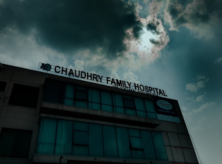 A hospital building with a sign that reads 'Chaudhry Family Hospital', positioned under a cloudy sky with visible sunlight peeking through. The building has multiple windows, some of which are covered or tinted.