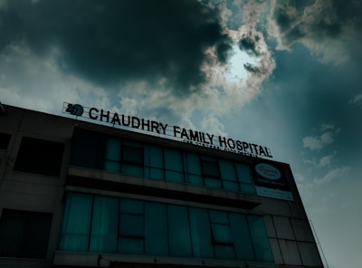 A hospital building with a sign that reads 'Chaudhry Family Hospital', positioned under a cloudy sky with visible sunlight peeking through. The building has multiple windows, some of which are covered or tinted.