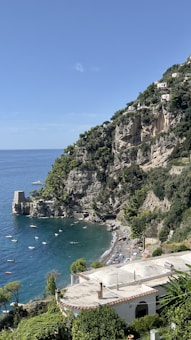 A scenic coastal view featuring rugged cliffs with greenery and multiple white buildings perched on the slopes. The clear blue sea is dotted with small boats, and a stone tower stands near the shoreline. A sandy beach is visible with sunbathers and umbrellas, surrounded by lush vegetation.