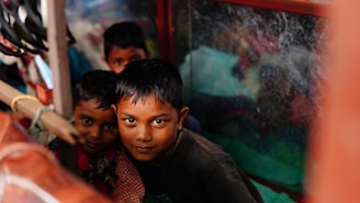 Three children are closely huddled together inside a small space, looking directly at the camera. The area is cluttered, with a window to the right partially obscured by dirt or scratches. The lighting is dim, casting shadows on their faces.