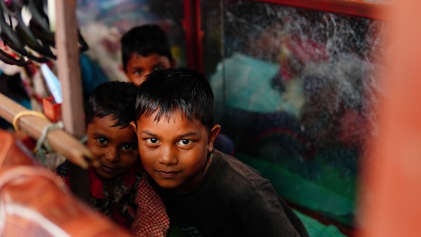 Three children are closely huddled together inside a small space, looking directly at the camera. The area is cluttered, with a window to the right partially obscured by dirt or scratches. The lighting is dim, casting shadows on their faces.