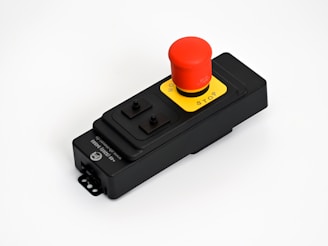 A black rectangular emergency stop button with a large red button labeled 'STOP' and 'ON'. The button is mounted on a black housing with a yellow background around the button. There are additional buttons or switches on the housing.