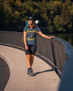 A woman wearing outdoor hiking gear stands on a curved pathway bordered by a railing. She is wearing a backpack and looking into the distance. The backdrop features dense green foliage, suggesting a park or natural setting.