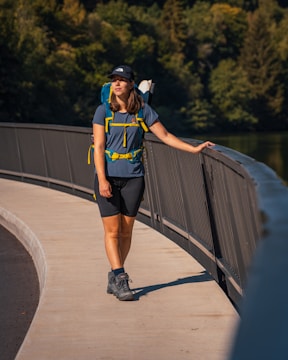 A woman wearing outdoor hiking gear stands on a curved pathway bordered by a railing. She is wearing a backpack and looking into the distance. The backdrop features dense green foliage, suggesting a park or natural setting.