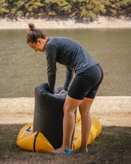 A person is outdoors, inflating or assembling a piece of equipment, likely a yellow inflatable raft or kayak, near a body of water. The individual is wearing a long-sleeve top and shorts, standing on grass with wooded area visible in the background.