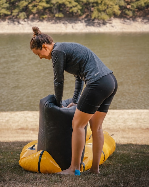 A person is outdoors, inflating or assembling a piece of equipment, likely a yellow inflatable raft or kayak, near a body of water. The individual is wearing a long-sleeve top and shorts, standing on grass with wooded area visible in the background.