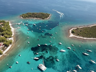 An aerial view of a coastal area with clear turquoise waters where numerous boats and yachts are scattered throughout the bay. Several small islands with dense greenery and rocky shores are visible. The scene suggests a popular recreational or tourist spot, with calm waters perfect for boating.