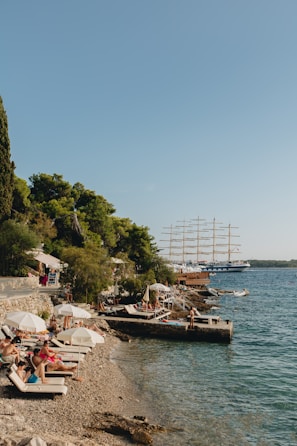 A scenic coastal area with a pebble beach lined with sunbeds and umbrellas, occupied by people relaxing under the sun. Lush green trees border the beach, and a large sailing ship with multiple masts is visible on the calm blue sea. The atmosphere suggests a leisurely seaside vacation.