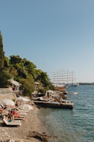 A scenic coastal area with a pebble beach lined with sunbeds and umbrellas, occupied by people relaxing under the sun. Lush green trees border the beach, and a large sailing ship with multiple masts is visible on the calm blue sea. The atmosphere suggests a leisurely seaside vacation.
