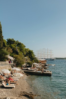 A scenic coastal area with a pebble beach lined with sunbeds and umbrellas, occupied by people relaxing under the sun. Lush green trees border the beach, and a large sailing ship with multiple masts is visible on the calm blue sea. The atmosphere suggests a leisurely seaside vacation.