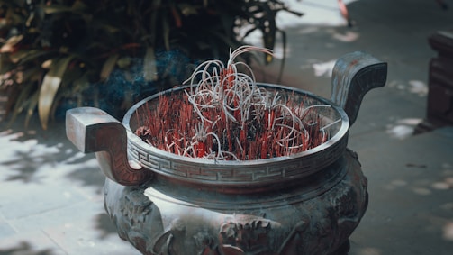 An ornate bronze incense burner filled with numerous red incense sticks, some of which are burnt down and curling over. Smoke from the incense creates a mystical atmosphere, while greenery in the background suggests an outdoor setting.